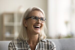 Smiling woman with straight, shoulder-length gray hair and black-rimmed glasses, wearing a plaid shirt, sits on a couch in a softly lit homey room with blurred shelves in the background.