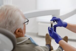 A dentist in purple gloves shows a dental implant model to an older man sitting in a dental chair, demonstrating the implant and artificial tooth in a clinical setting.